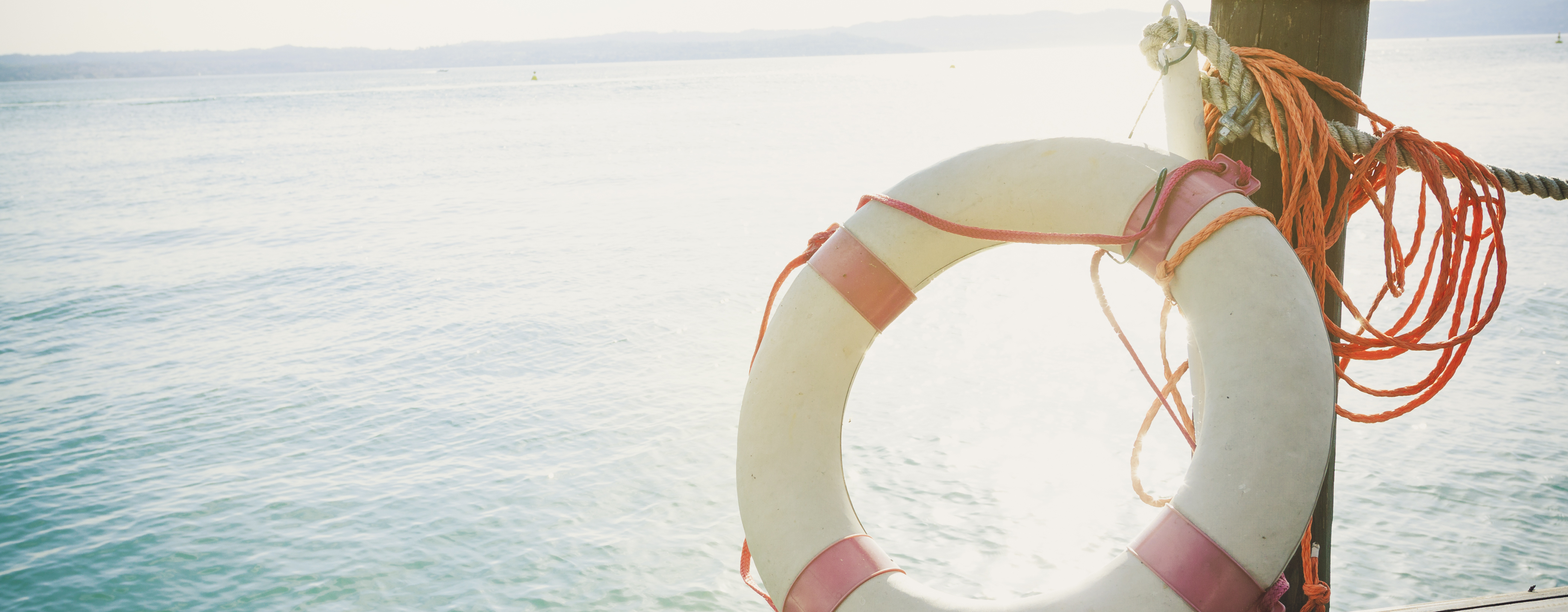 A traditional red and white life preserver is resting against a pole on a wooden dock, overlooking green waters. The sun is shining on everything, washing the color out. In the distance there are some hills on the skyline. The life preserver is connected to the dock rope with thin, orange rope | Snag-A-Slip News & Updates